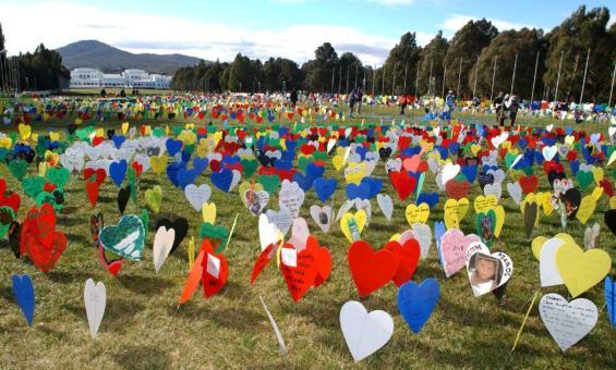 A large field filled with colorful heart-shaped signs, some with messages and images, displayed on the grass. Old Parliament House is visible in the background.