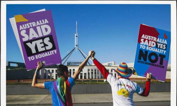 A photo of 2 people with their backs to the camera, facing Australia's Parliament House, holding hands, lifting them high. Each is holding a large sign in their outside hands that reads 'Australia said Yes to equality' and 'Australia said Yes to equality, not discrimination'.