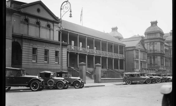 black and white photo of old buildings with old cars in the street out the front