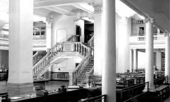 Black and white photo from inside the National Bank head office. 2 ornate staircases in the middle of the image surrounded by white pillars and desks. 