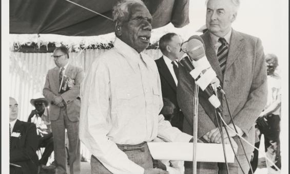 Vincent Lingiari addresses a crowd at a microphone while Gough Whitlam stands beside him. Several people are visible in the background, observing.