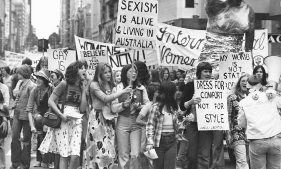 A group of women marches down a city street, holding various protest signs. Signs include messages like "Sexism is alive & living in Australia" and "Dress for comfort, not for style." A large sculpture is visible in the background.