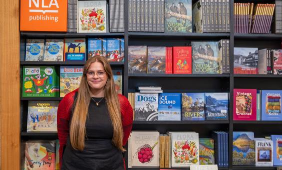 Young woman, Maddie from NLA Publishing, standing in front of a wall of NLA Publishing books in the Bookshop