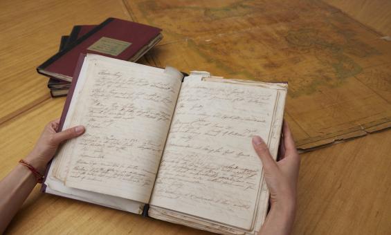 Hand holding an old notebook with handwritten text. In the background is a stack of books and an old map.