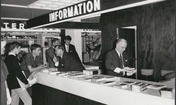 A black and white photograph of a group of men standing at a white information desk. Above the desk is a large sign that says INFORMATION. Men behind the counter and speaking and hanging out brochures.