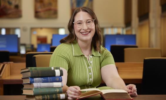 Zoe Smith, an NLA Scholar, sitting at a table in the Main Reading Room with a pile of books and smiling
