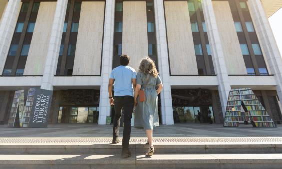 Man and woman in casual wear holding hands and laughing as they walk up the stairs towards the National Library entrance