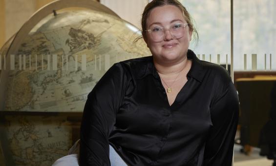 Woman, Jacinta Mackay, sitting in front of a window looking into the Main Reading Room with a large globe on the other side