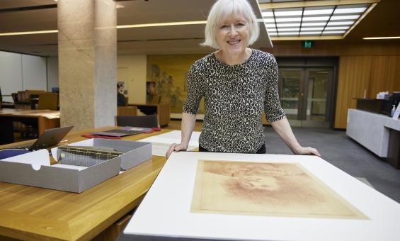 Woman, Dr Christina Thompson, standing in the Special Collections Reading Room, leaning on a table with a large portrait on it and smiling