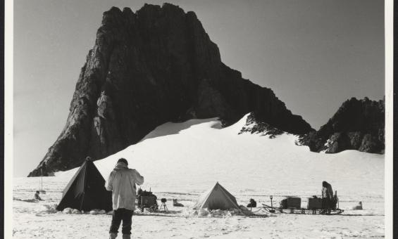 A man standing on a snowy plain, in front of tents, sledges with scientific equipment, scientists and sled dogs. A large rocky outcrop looms in the background.