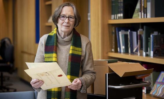 A woman with short hair and glasses smiles and looks at the camera while she holds a folded document in her hands. In the background are bookshelves with books and an open box on a trolley.
