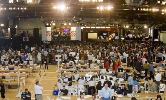 People standing, sitting at desks, and filming with news cameras in a brightly lit room