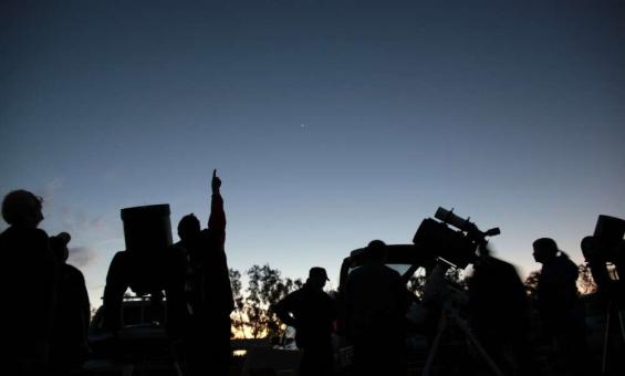 A group of people at dusk standing beside small telescopes and pointing at the sky.