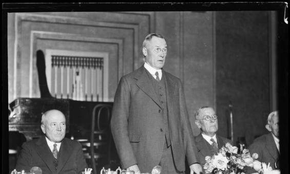 A gentleman standing behind a table in a suit addressing the room 