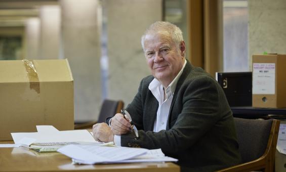 Man, Professor Robin Gerster, smiling and sitting at a table in the Special Collections Reading Room with manuscipt material in front of him