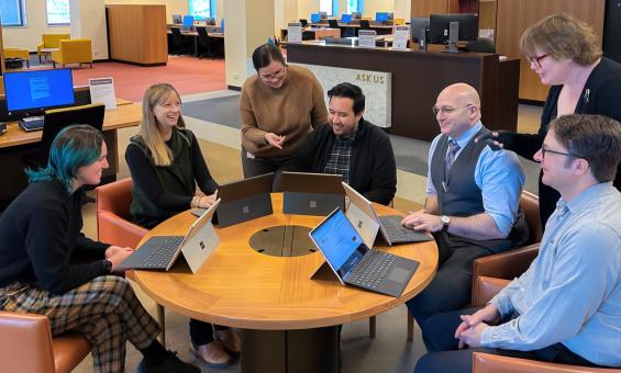 Seven librarians of the National Library sitting or standing around a table talking, smiling and looking at laptops