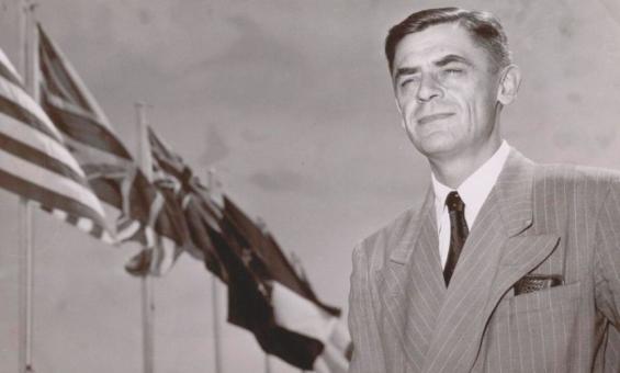 A black and white photograph of a man in a suit standing in front of flagpoles with the US and Australian flags