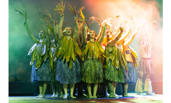 Group of dancers wearing large leaves around their necks and grass skirts, standing in a circle with their arms raised