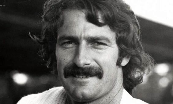 A black and white portrait of Dennis Lillee wearing cricket whites. A jumper is draped over his right shoulder.