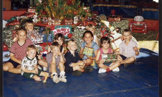 Group of 10 young children sitting under a Christmas tree with presents