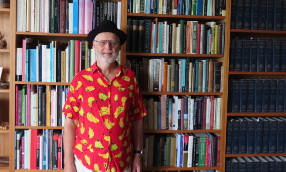 Professor Matthew Spriggs is standing in front wooden bookshelves filled with books. He is wearing glasses, a red short-sleeve shirt patterned with yellow bananas and a black hat.