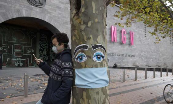 A man wearing a face mask leaning against a tree that is decorated with a face mask and eyes outside the NGV in Melbourne