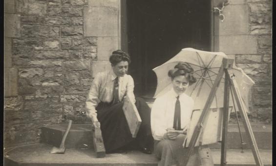 A black and white image of two women sitting on concrete steps, one holding woodworking tools, the other painting at an easel and holding a parasol