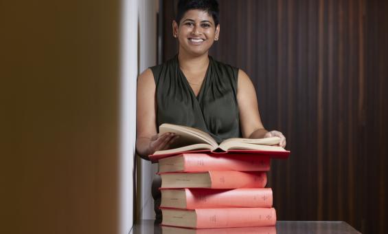 Natasha Naidu standing at a desk behind a stack of books holding one open and smiling at the camera