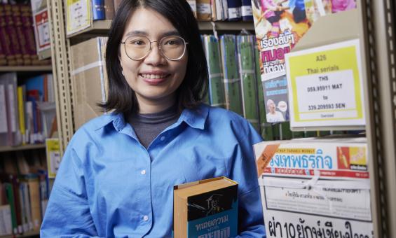 Woman with dark hair, Suprawee (Earn) Asanasak, standing in the stacks of the Library smiling and holding a book