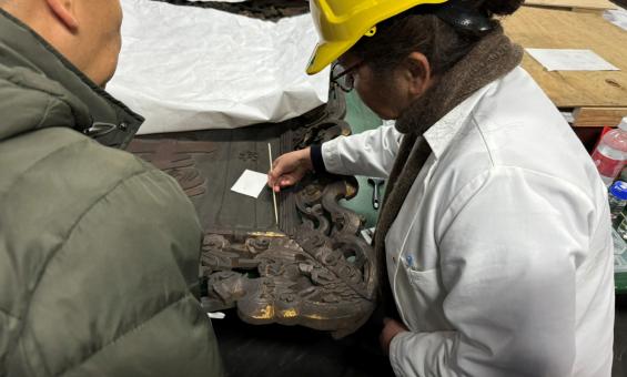 A woman stands on the right-hand side of this image holding a cleaning tool and applying it to a plaque. A man is on the left-hand side watching the cleaning process. 