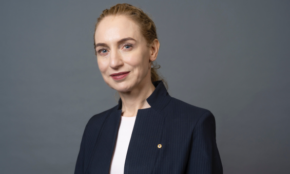 A woman with long hair tied back stands in the centre of the image wearing a white shirt and navy jacket