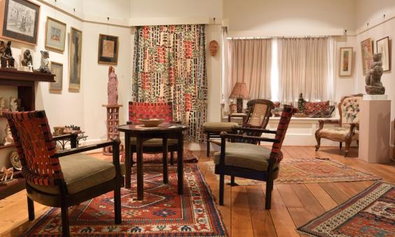 A cosy living room. A small table surrounded by four chairs takes up much of the image. There are a few other seats in the image, and artwork and sculpture adorning the walls and shelves lining the walls.