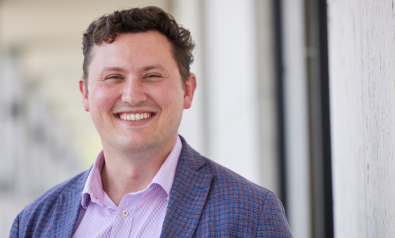 A man wearing a lilac button down shirt and a blue and red checkered blazer while smiling at the camera