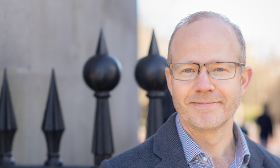 A man wearing glasses, blue shirt and jacket, standing in front of a wrought iron fence