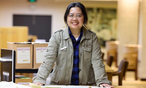 A woman wearing a grey jacket stands leaning on a table with papers in a library setting