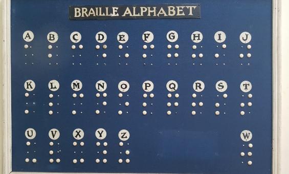 A braille alphabet chart. Each letter of the alphabet is in a large white circle with the corresponding braille code for that letter below it. The background of the chart is blue. 