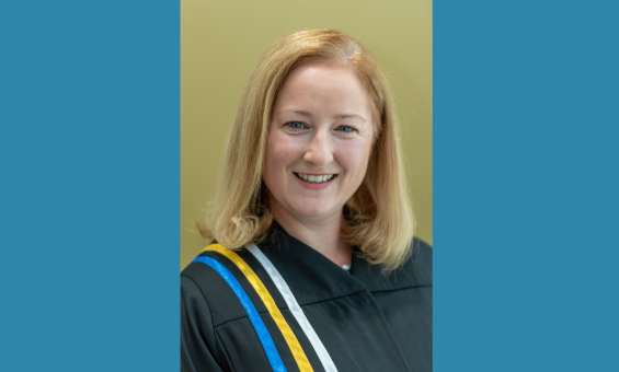 A blond-haired woman wearing courtroom robes with blue, yellow and white ribbons on the right shoulder