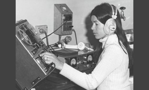 A black and white photo of a woman wearing headphones, operating a vintage audio recording or mixing console in a studio setting.