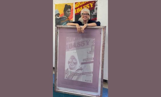 A woman with blonde hair and glasses leaning on a purple portrait in a studio with some Indigenous posters behind her.