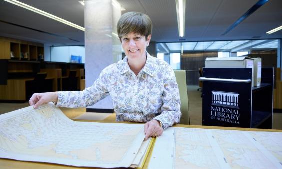 A smiling woman with short brown hair sits at a desk, holding open a large, detailed historical map. She wears a floral-patterned blouse and looks directly at the camera..