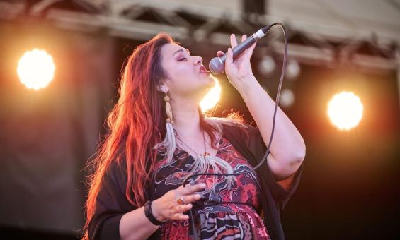 A female singer with long, reddish-brown hair performing live on stage under bright spotlights. She is holding a microphone and appears to be singing passionately.