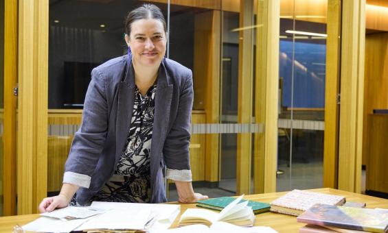 A woman with dark hair pulled back smiles at the camera while leaning on a wooden table. She wears a grey blazer over a patterned blouse. Several books and open notebooks are spread across the table in front of her.