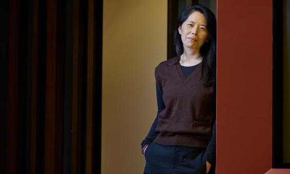 A woman with long dark hair stands leaning against a wall, smiling gently at the camera. She wears a brown V-neck sweater over a black shirt and dark pants, with her hands in her pockets.