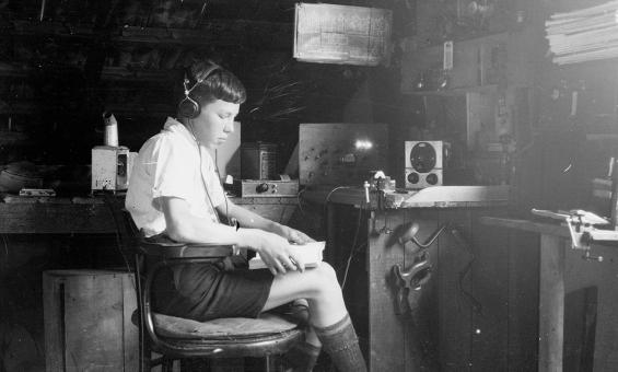 A young person wearing headphones sits at a cluttered workbench in a dimly lit, rustic room, focused on operating vintage radio equipment. The scene suggests a hobbyist or amateur radio operator in a makeshift workshop filled with tools, electronic devices, and schematic diagrams.